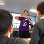 Picture of Rosie pouring a small tube of yellow liquid into a larger tube with a layer of yellow liquid and a layer of clear liquid.Two primary students are watching.